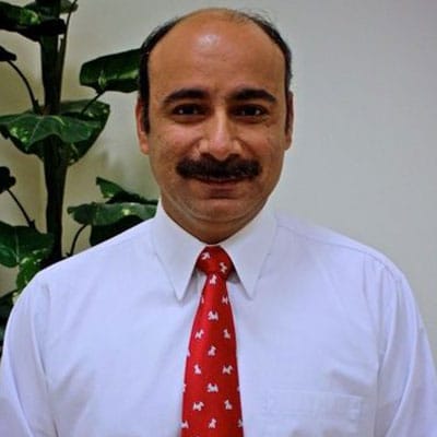 A man with a mustache and receding hairline, wearing a white dress shirt and a bright red tie with white dog patterns, is standing in front of a leafy green plant. The veterinarian is smiling at the camera.