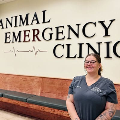 A smiling veterinary professional stands in front of a wall sign that reads "Animal Emergency Clinic" with a heart rate graphic; there are empty benches along the wall.