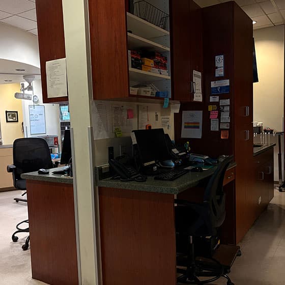 A hospital or clinic nurse station with two desks, computers, office chairs, telephones, and shelves stocked with supplies. The area is tidy and well-organized, with notes and charts posted on the cabinets.