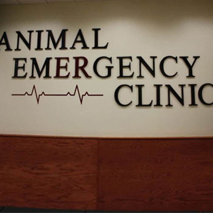 home_s4_1 Wall signage reading "Animal Emergency Clinic" with a heartbeat line graphic. The word "Emergency" stands out in red while the other words are in black. The background is a light cream color, and the lower part of the wall has a wooden panel, giving it a professional vet clinic atmosphere.