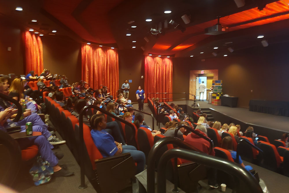 A large lecture hall with red seats and several people sitting, mostly looking at their phones. The room has dark walls and is dimly lit. A veterinarian at the front appears to be speaking or presenting.