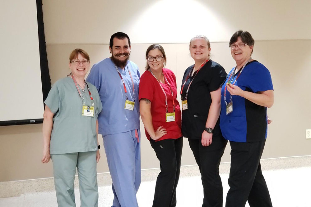 Five healthcare professionals, including a veterinarian, stand together and smile in a well-lit room. They are dressed in colorful scrubs: one in light green, one in light blue, one in red, one in black, and one in dark blue. Each person has a badge and a lanyard around their neck.