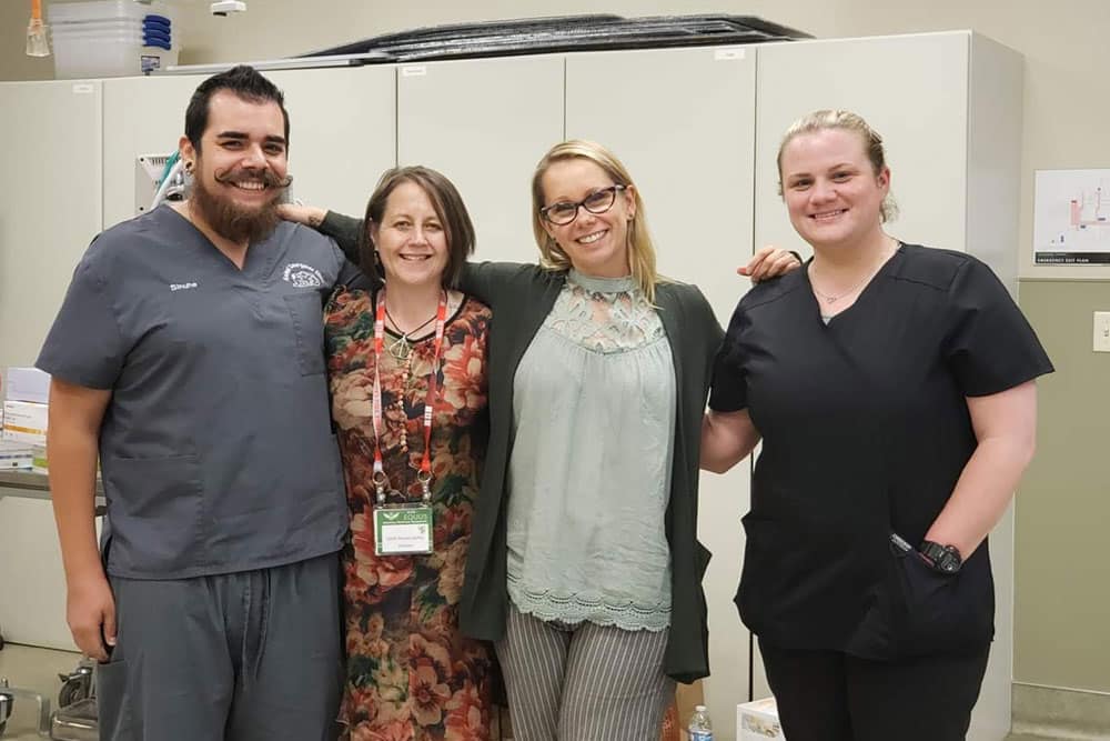 Four people stand together in a room with cabinets. They are smiling and have their arms around each other’s shoulders. One man, possibly a veterinarian, is wearing scrubs, one woman is in a floral dress, another woman is in a blouse with a cardigan, and the last woman is in black scrubs.