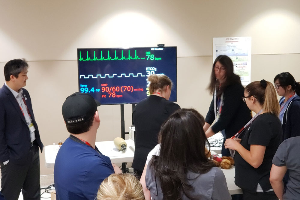 A group of people, some in medical attire, gather around a medical simulation setup. A veterinarian is adjusting equipment while a monitor displays vital signs: SpO2 99.4%, ECG 90/60 (70) mmHg, heart rate 78 bpm. A poster is seen on the wall in the background.