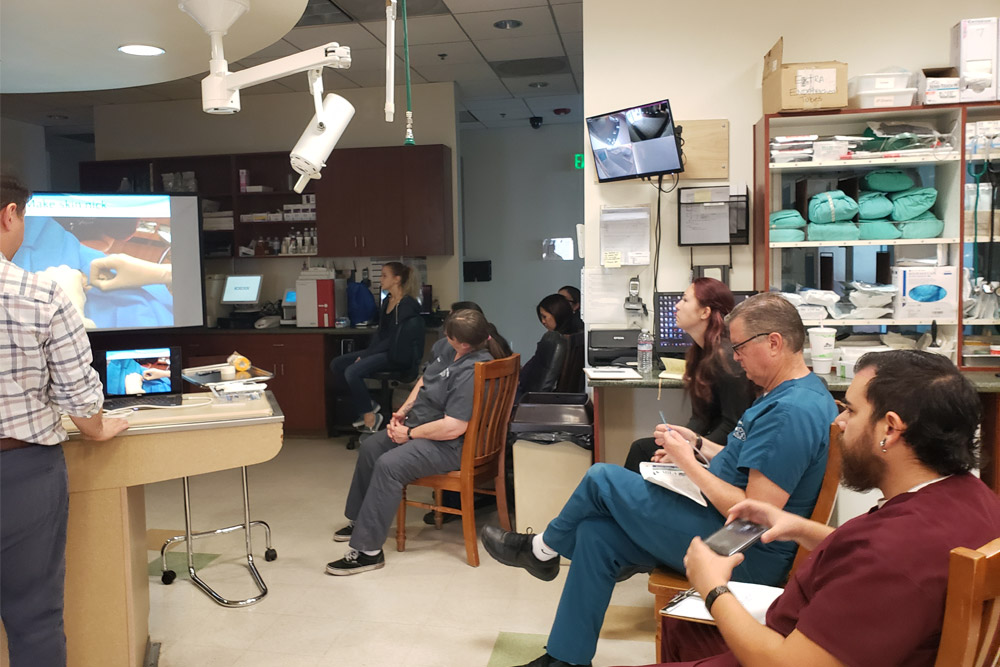 A group of medical professionals, including a veterinarian, is gathered in a hospital room for a training session. They are watching a presentation displayed on a TV screen. Various medical supplies and equipment are visible in the background. Some are seated, while others stand and observe.