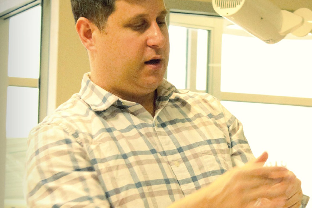 A man with short dark hair, wearing a plaid shirt, is gesturing with his hands. He appears to be explaining something or engaging in a discussion, possibly about his work as a veterinarian. Behind him are large windows, and there is a white ceiling-mounted projector in the upper corner of the image.