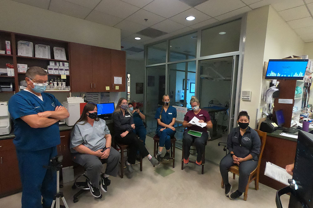 A group of healthcare professionals, including a vet in scrubs and masks, gather in a hospital treatment room. Some are seated while others stand, listening attentively to someone out of frame leading the discussion. Medical supplies and equipment are visible in the background.