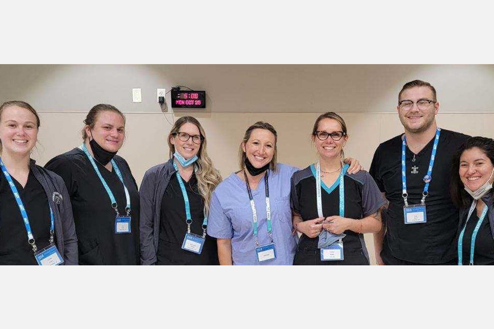 A group of seven people, wearing ID badges and medical uniforms, stand together smiling. Some have stethoscopes around their necks. They are in a well-lit indoor setting with a clock showing 10:25. One person in the center wears a light blue scrub top that hints at their role as a veterinarian.