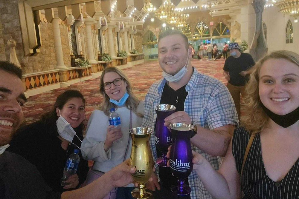 A group of five people, including a veterinarian, stand indoors, smiling and holding up drinks in celebratory goblets. Some are carrying bottled water, and two are wearing face masks under their chins. The background features ornate columns and bright lights, suggesting a festive environment.