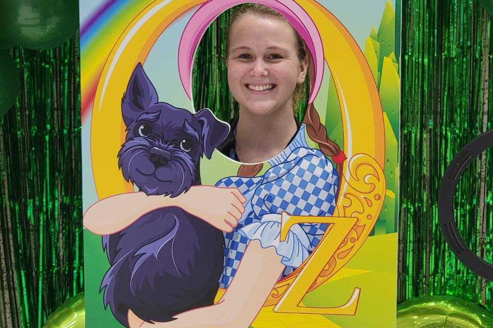 A veterinarian smiles through a cutout standee of Dorothy from "The Wizard of Oz," hugging Toto. The colorful background features a rainbow and green scenery, with green metallic streamers as a backdrop. The vet wears a blue and white checkered pattern similar to Dorothy's dress.