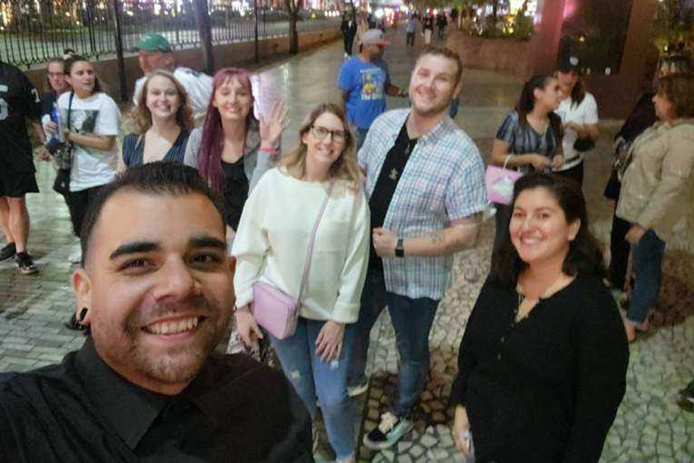 A group of six people, including a veterinarian, smile and pose for a photo on a tiled sidewalk at night. The person in the foreground is taking a selfie with others behind them. They appear happy and are standing in an urban area with other people in the background.