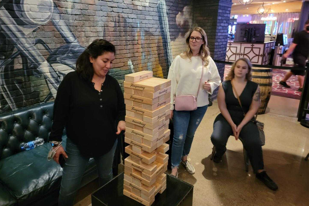 Four women, including a veterinarian, are engaged in a game of Giant Jenga in an indoor setting with brick walls and artwork in the background. One woman is carefully pulling out a block while the others watch. There is a green couch, a water bottle, and a bar counter in the scene.