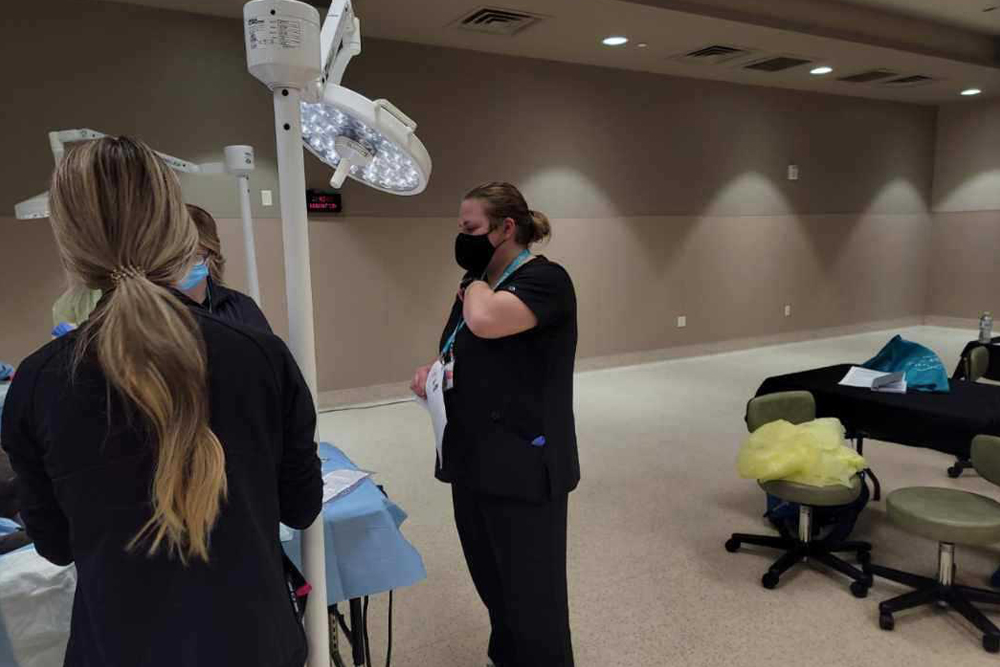 A medical professional, resembling a veterinarian, wears a black face mask while standing in a hospital room. They are examining a patient or discussing with colleagues who are partly visible. Surgical lights illuminate the space, and various medical equipment and furniture are placed around the room.
