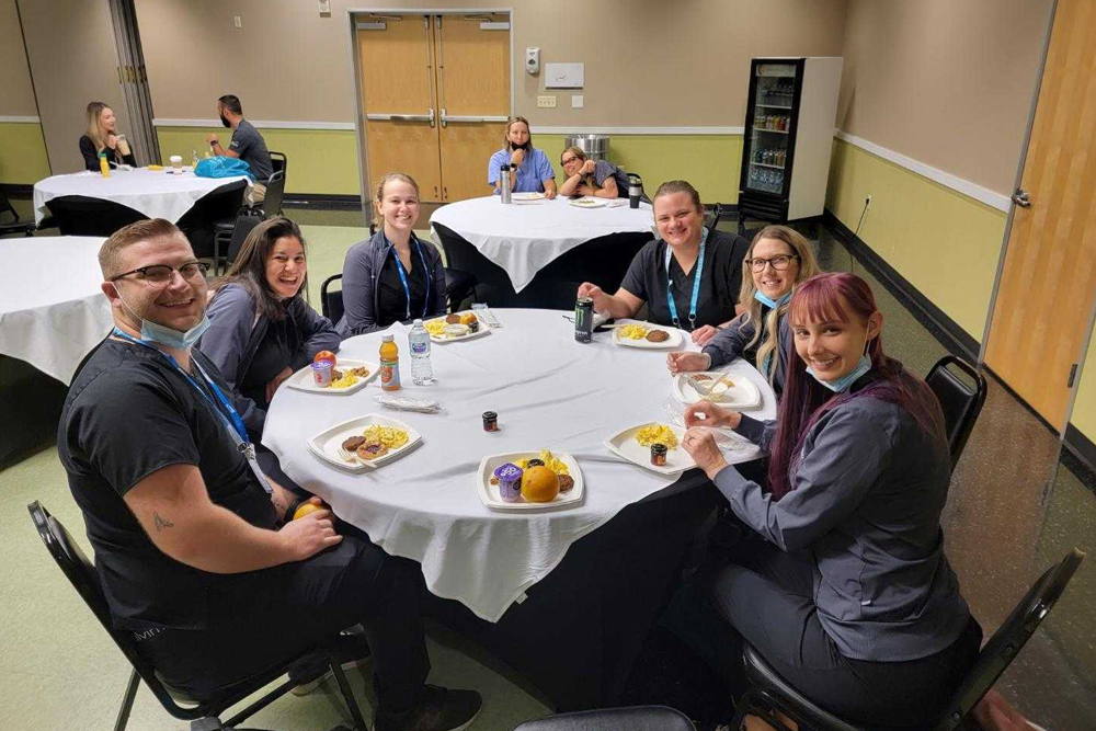 Seven people, including a veterinarian, sit around a table in a room, sharing a meal. The table is covered with a white tablecloth, and plates of food and drinks are in front of them. Additional tables with more people are seen in the background. The atmosphere appears friendly and relaxed.