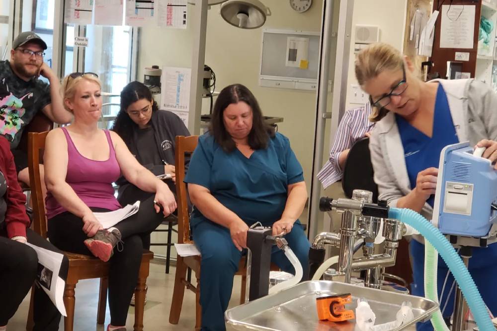 A group of people in a medical setting attentively watch a woman in scrubs, possibly a veterinarian, demonstrating a piece of medical equipment. Some individuals are seated, holding papers with focused expressions, while the woman in scrubs operates a device beside a metal sink and various medical instruments.