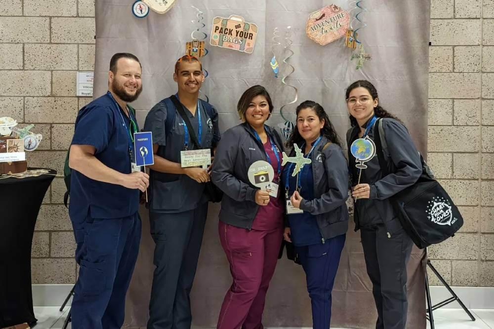 Five people dressed in medical scrubs, likely veterinarians, stand in front of a travel-themed backdrop holding various travel-related props. The backdrop includes images of luggage and travel signs. They are smiling and appear to be at an event or gathering.