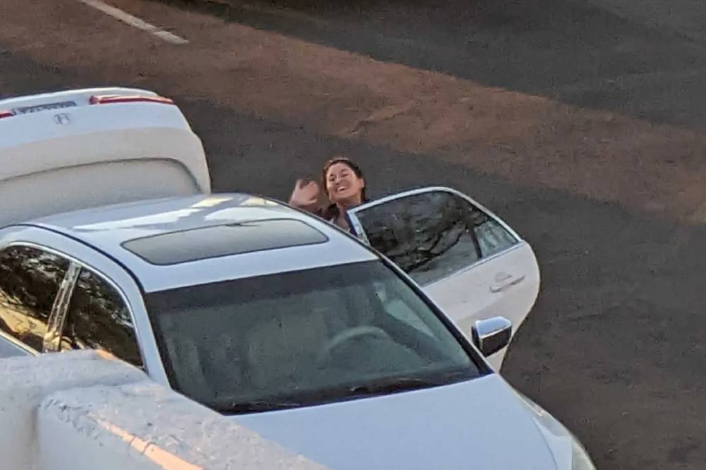 A woman standing next to a white car waves with a smile, possibly looking for her next appointment as a veterinarian. The car is parked on a paved surface, and the trunk of the car is open. The scene appears to be captured from above.