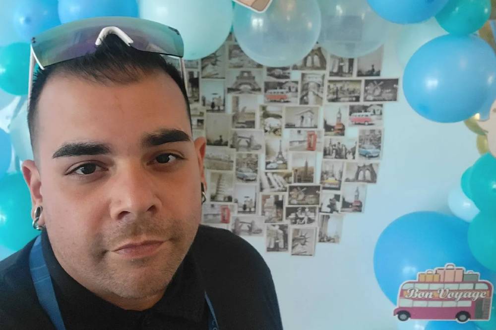 A veterinarian with short dark hair and sunglasses on their head is standing in front of a blue and white balloon decoration. Behind them is a collage of various travel-themed photographs and a "Bon Voyage" sign featuring a car with luggage on top.