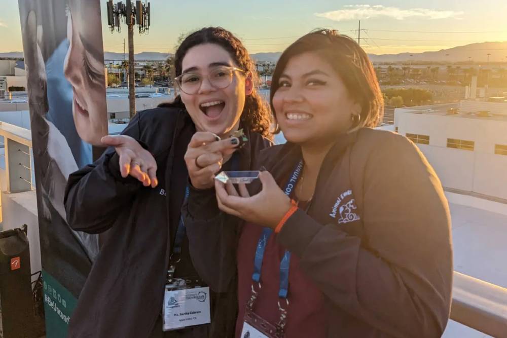 Two smiling women are enjoying dessert outdoors on a sunny day. One woman, a veterinarian, is holding a small dessert bowl in one hand and a spoon in the other, while the second woman holds the bowl from the opposite side. Both are wearing lanyards and jackets. A cityscape is visible in the background.