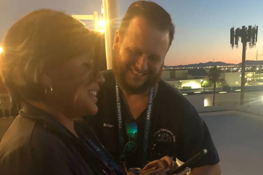 Two individuals wearing lanyards, likely veterinarians attending a conference, laugh and smile while looking at a smartphone. They are outdoors at dusk, with a cityscape and a lit tower visible in the background. The vets seem to be enjoying a joyful moment amidst the event's activities.