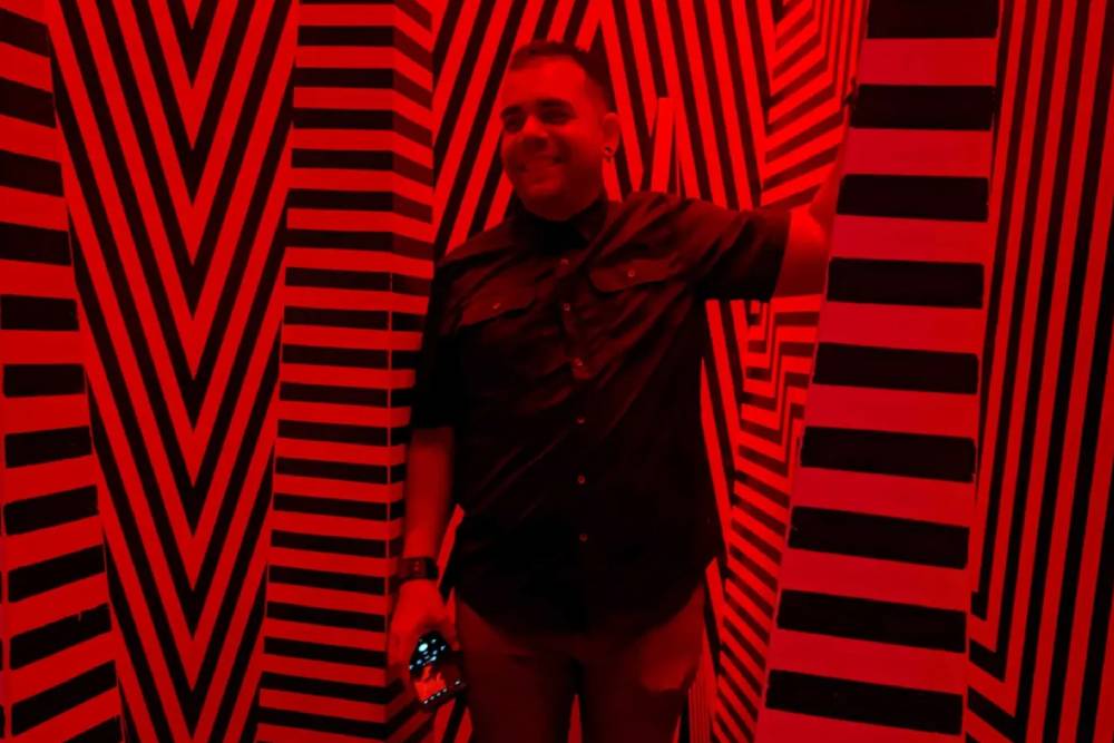 A person smiles while standing in a room with intense red lighting and walls covered with black and white zig-zag and vertical stripes. The veterinarian, holding a camera, wears a dark shirt and pants.