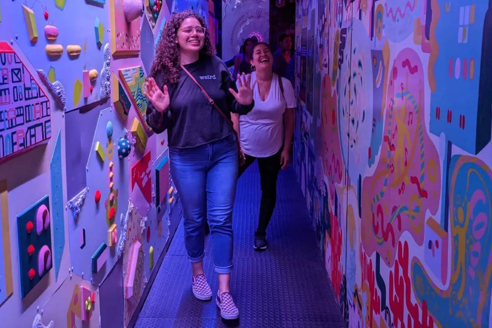 Two women walk through a colorful, art-filled hallway. The woman in front, a veterinarian wearing a black hoodie and jeans, smiles with her hands raised. The woman behind her, wearing a white shirt and black pants, follows while smiling. The walls are decorated with vibrant, abstract artwork.