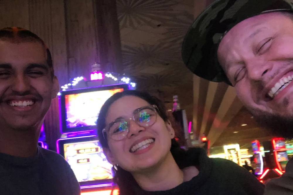 Three people, including a veterinarian, are smiling and taking a selfie in a brightly lit room filled with slot machines, likely a casino. The background features neon lights and digital displays. The mood appears joyful and vibrant.