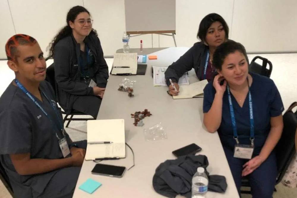 Four people in medical scrubs sit around a table with notebooks, pens, a phone, water bottle, and small snacks. They are smiling and appear to be in a discussion or study session. The room is brightly lit with a plain background. Among them is a veterinarian sharing insights from their practice.
