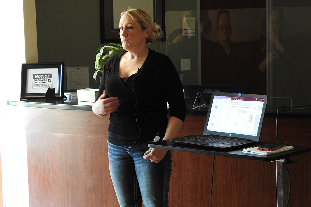 A woman wearing a black top and jeans stands in front of a desk, giving a presentation. She is speaking and gesturing with her hand. A laptop displaying her veterinarian research is on a small table to her right. A framed notice and plants are visible in the background.
