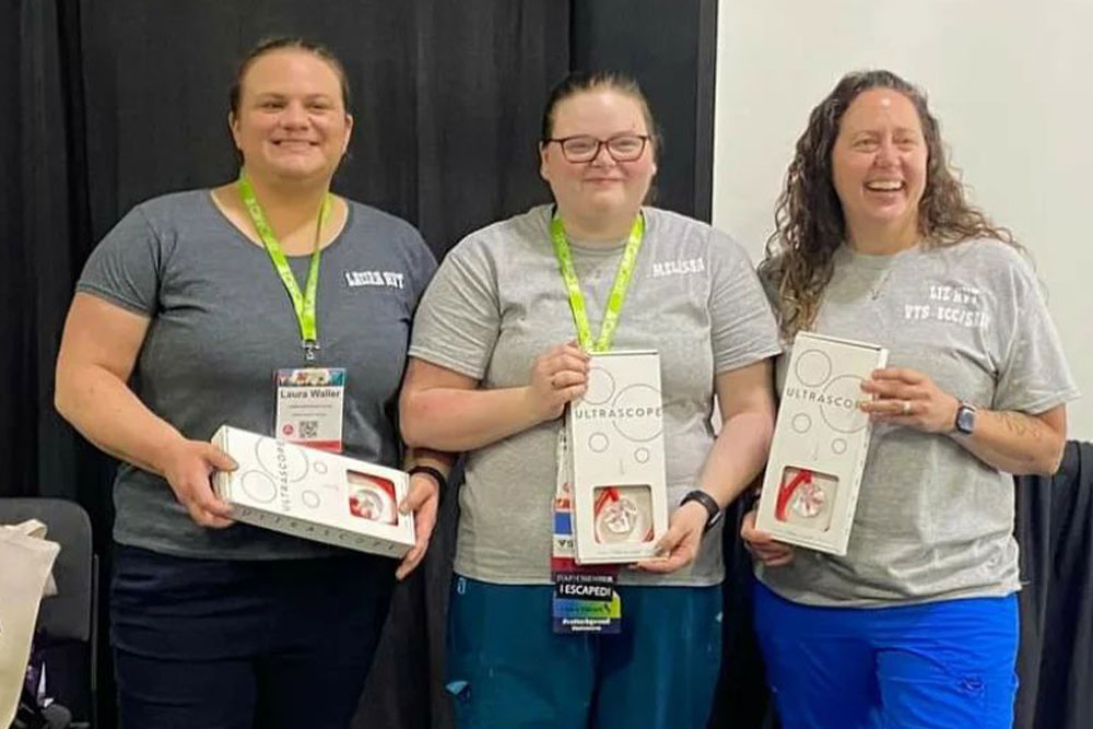 Three people standing and smiling, each holding an Ultrascope box. They are wearing grey shirts, and two are wearing green lanyards with name tags while one has a blue lanyard. They appear to be veterinarians at an event or conference, standing in front of a dark backdrop.