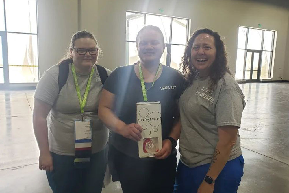 Three people are smiling indoors. The person in the middle, a veterinarian, holds a box labeled "Ultrascope." They all wear lanyards and casual clothing. The well-lit room has large windows in the background.