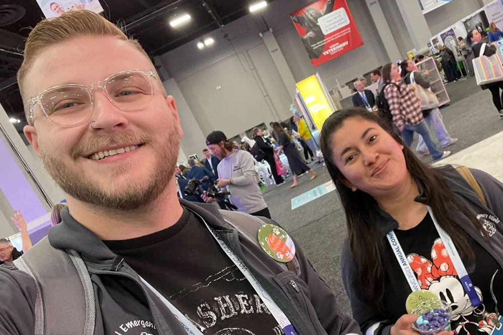 A man with glasses and a beard and a woman with long dark hair are smiling at a busy indoor event. The woman, wearing a Mickey Mouse shirt, is holding a toy. Both have lanyards around their necks. The man, who is clearly a veterinarian, seems to be enjoying the lively atmosphere filled with people and booths in the background.