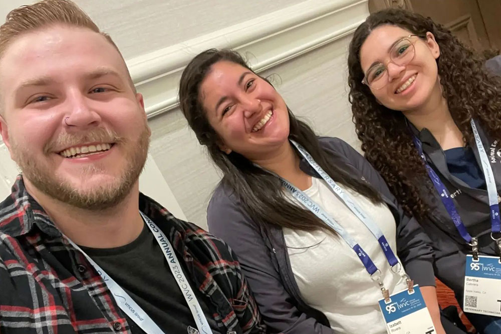 Three people are smiling and sitting together, wearing name tags and lanyards with "AWS" and "97th Annual" printed on them. One person, a veterinarian in a plaid shirt, sits beside another in a white top and gray jacket. The third, dressed in dark clothing with curly hair, completes the cheerful trio.