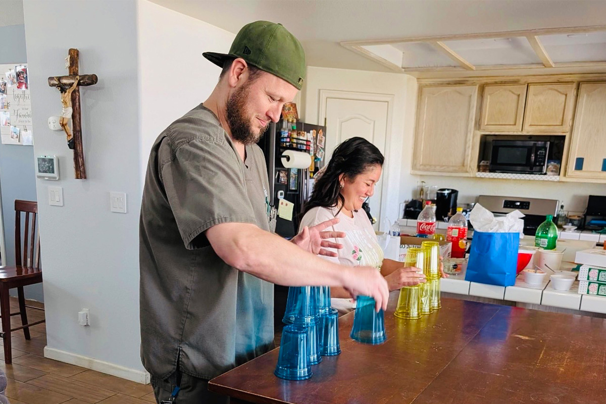 staff-event-1 A man and woman smile as they stack colorful plastic cups on a wooden table in a kitchen. The kitchen has white cabinets, a cross on the wall, and drinks and food on the counter in the background.