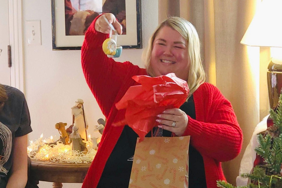 staff-event-10 A woman in a red cardigan smiles while holding up a small ornament and red tissue paper, standing next to a Christmas tree and holiday decorations, with a gift bag in her other hand.