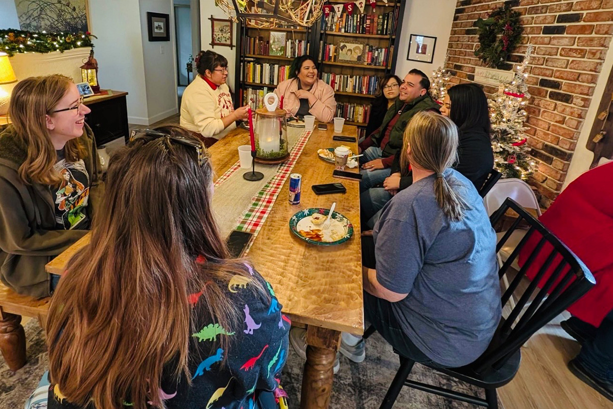 staff-event-11 A group of people sit around a wooden table in a cozy, festive living room decorated with a Christmas tree and holiday decor. They are talking, eating, and enjoying each other's company.