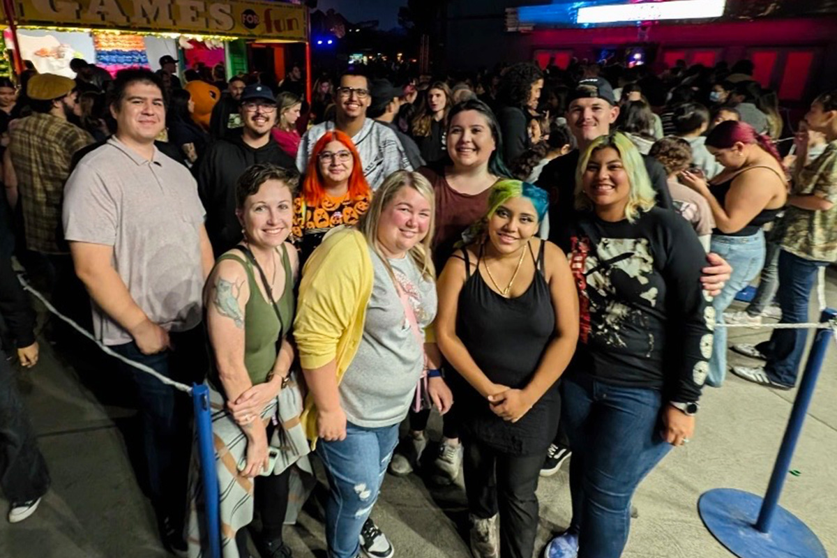 staff-event-13 A group of people smiling and posing for a photo outdoors at night, standing near a rope barrier with a crowd and colorful lights from a games booth in the background.