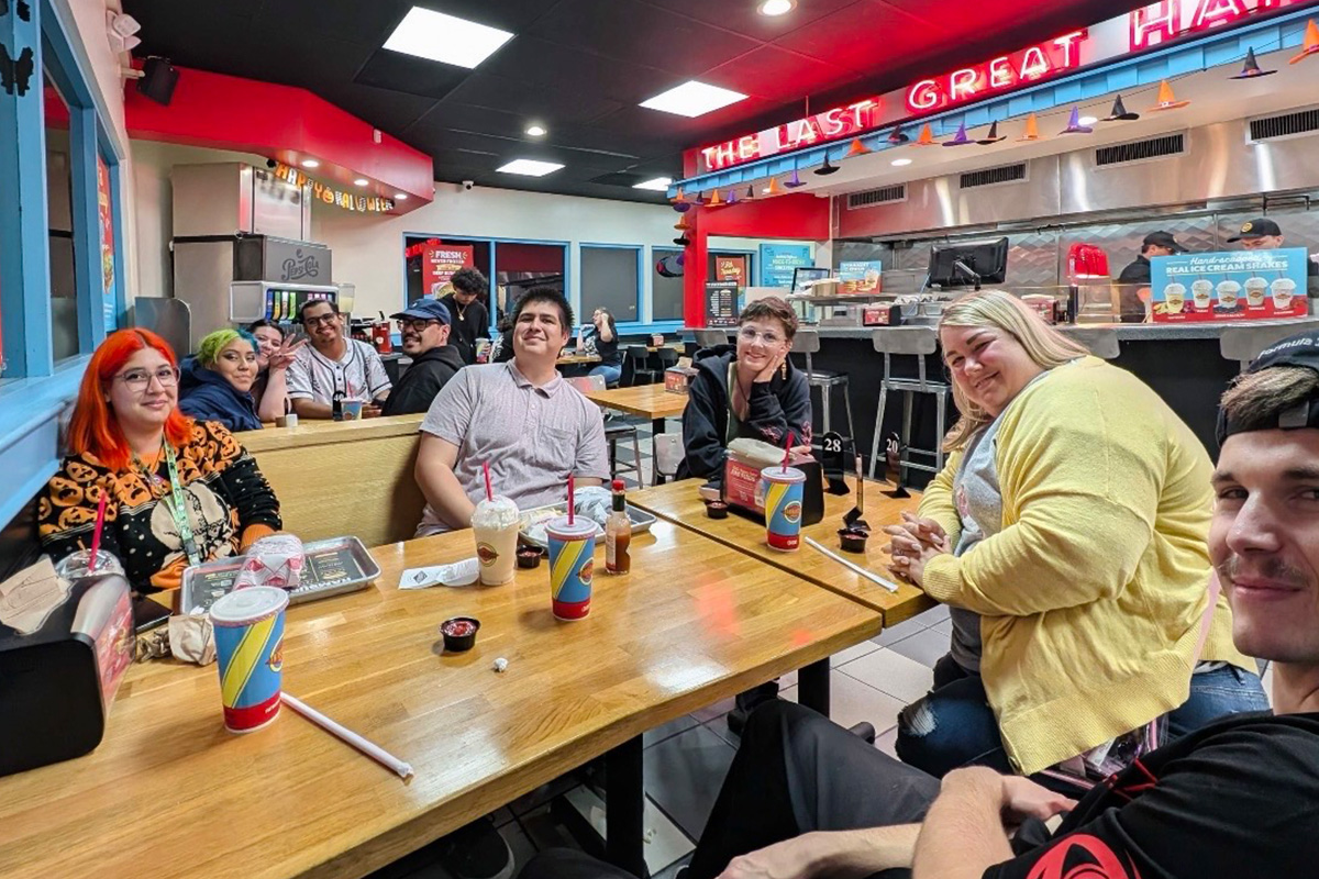 staff-event-14 A group of people sit at two connected tables in a brightly lit fast-food restaurant, smiling at the camera. They have drinks, trays, and food in front of them. The restaurant has red accents and a neon sign overhead.