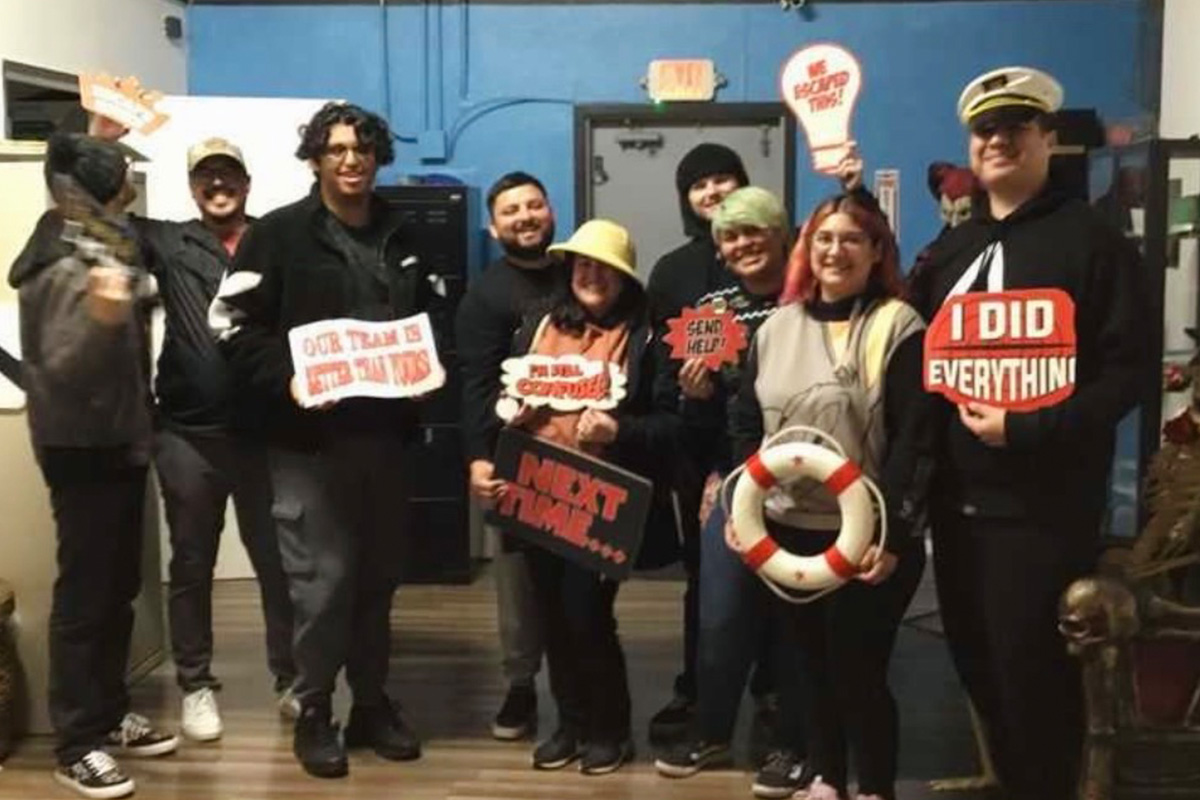 staff-event-15 A group of nine people stand indoors, smiling and holding themed signs and props like hats, a captain’s hat, and a life preserver. The signs include phrases like “I did everything!” and “Send help!” The setting appears playful and casual.