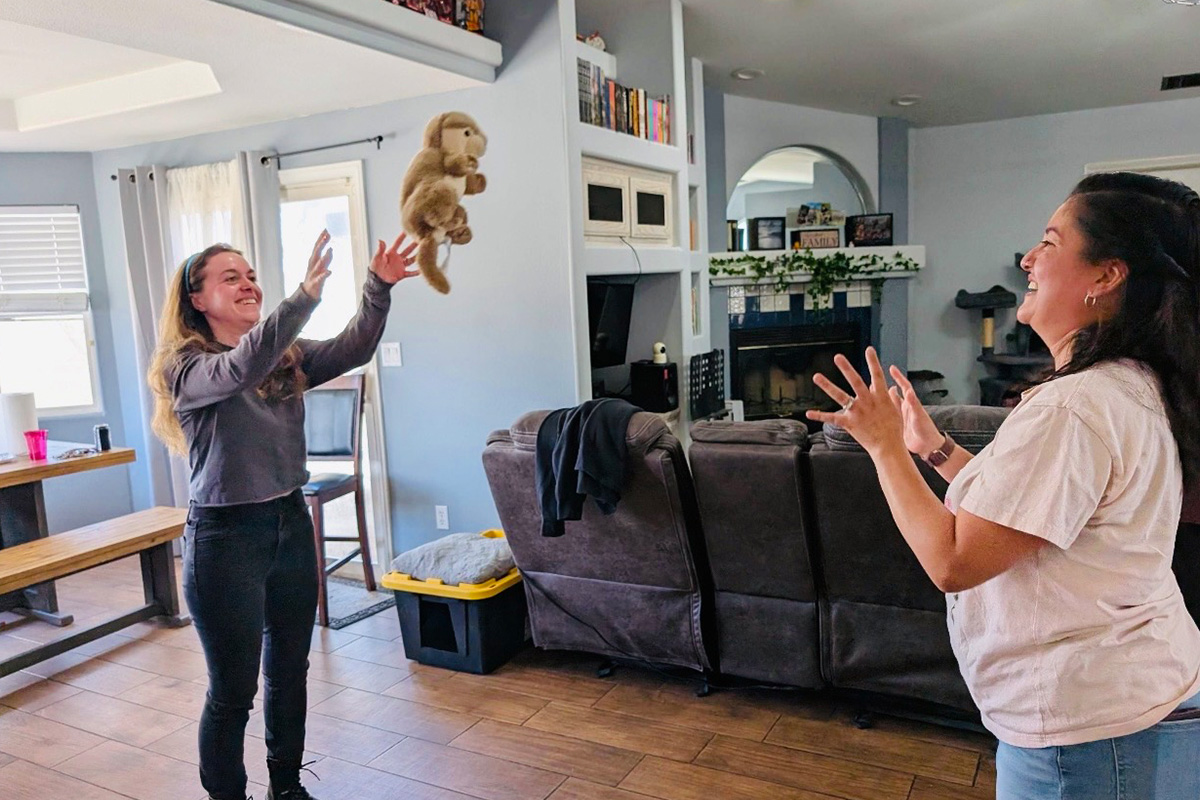 staff-event-2 Two women stand in a living room smiling and tossing a stuffed monkey toy to each other. The room has a couch, a dining table, and light streaming in from the windows.