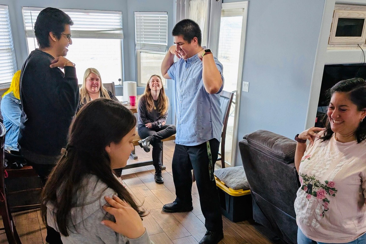 staff-event-3 A group of young adults gathers in a living room. Four are standing, smiling, and touching their shoulders, while three sit on chairs and a couch, watching and laughing. The mood is cheerful and relaxed.