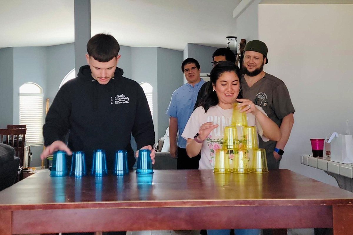 staff-event-5 Two people compete stacking plastic cups on a table, one with blue cups and the other with yellow cups. Three others stand behind them watching and smiling in a brightly lit room.