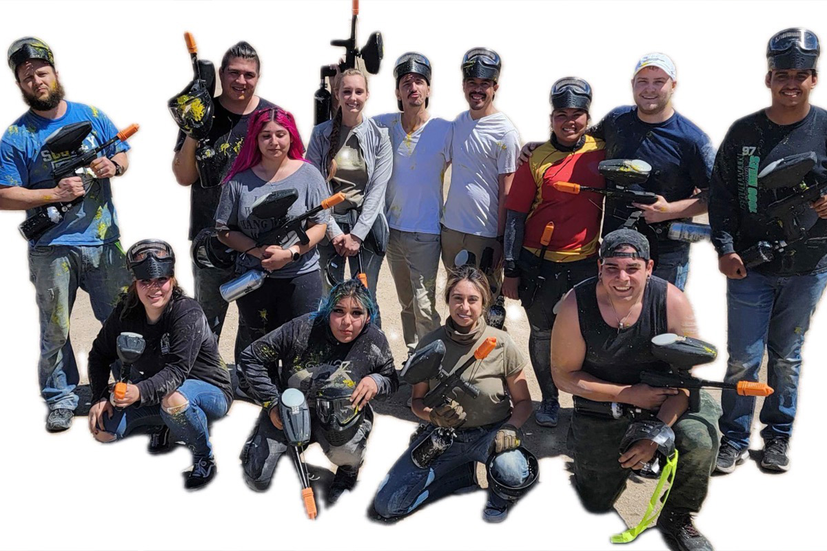 staff-event-6 A group of thirteen people pose together outdoors, wearing paintball gear and holding paintball guns. They are smiling and appear to have just played a game, with some visible paint splatters on their clothes.