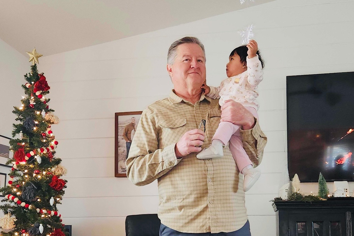 staff-event-7 A man holds a young child as they decorate a Christmas tree in a cozy living room. The tree is adorned with red and white ornaments, and festive decorations are visible on shelves and a fireplace.
