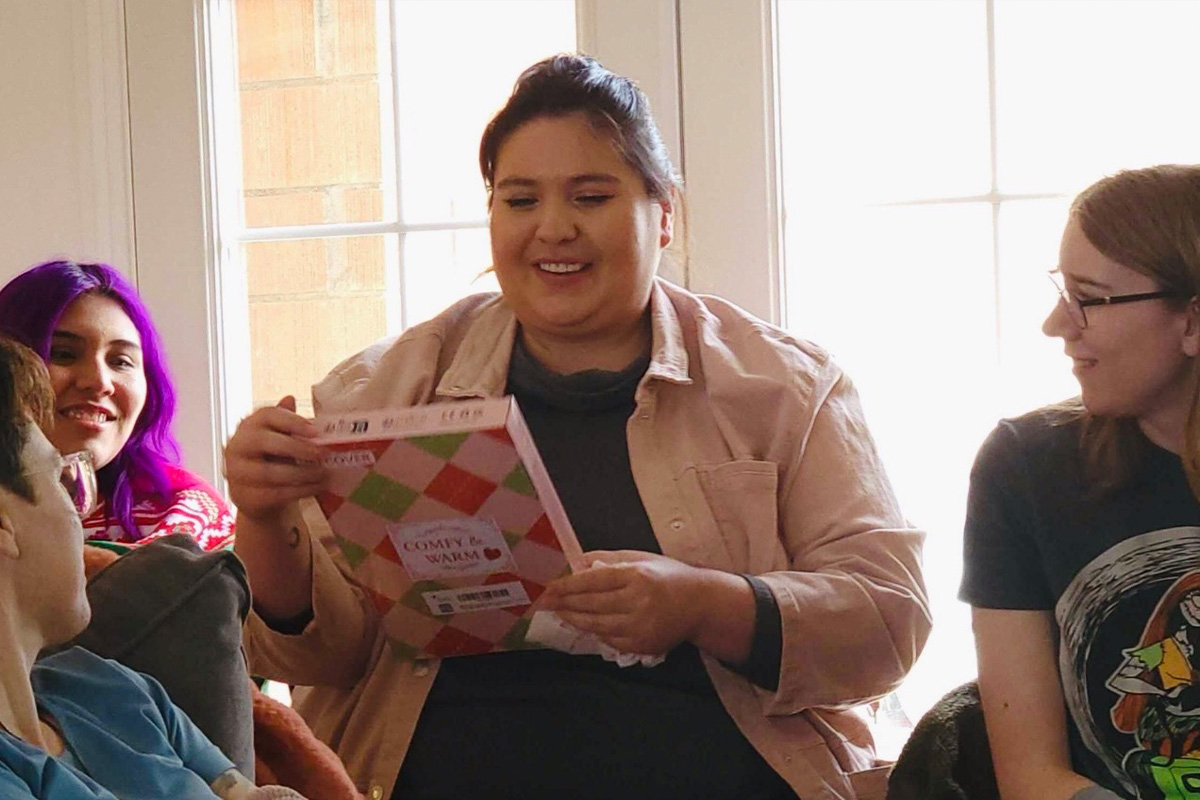 staff-event-8 A woman smiling and holding a cookbook surrounded by friends, sitting together indoors with sunlight streaming through large windows in the background.