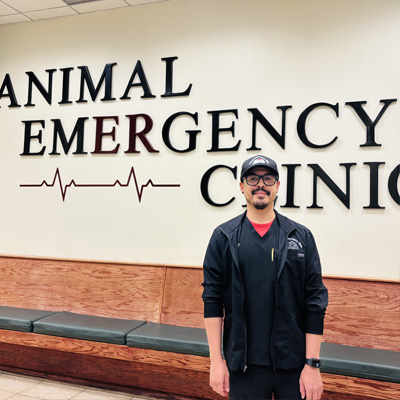 A man wearing glasses, a cap, and black scrubs stands in front of a wall sign that reads "Animal Emergency Clinic" with a red heartbeat line graphic. Benches line the wall beneath the sign.