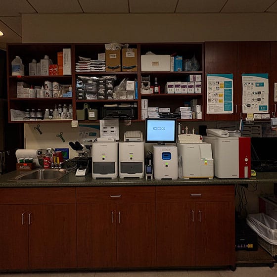 A laboratory workspace with medical equipment and machines on a counter, shelves above filled with supplies, bottles, boxes, and organized containers. Posters and instructions are posted on the wall.