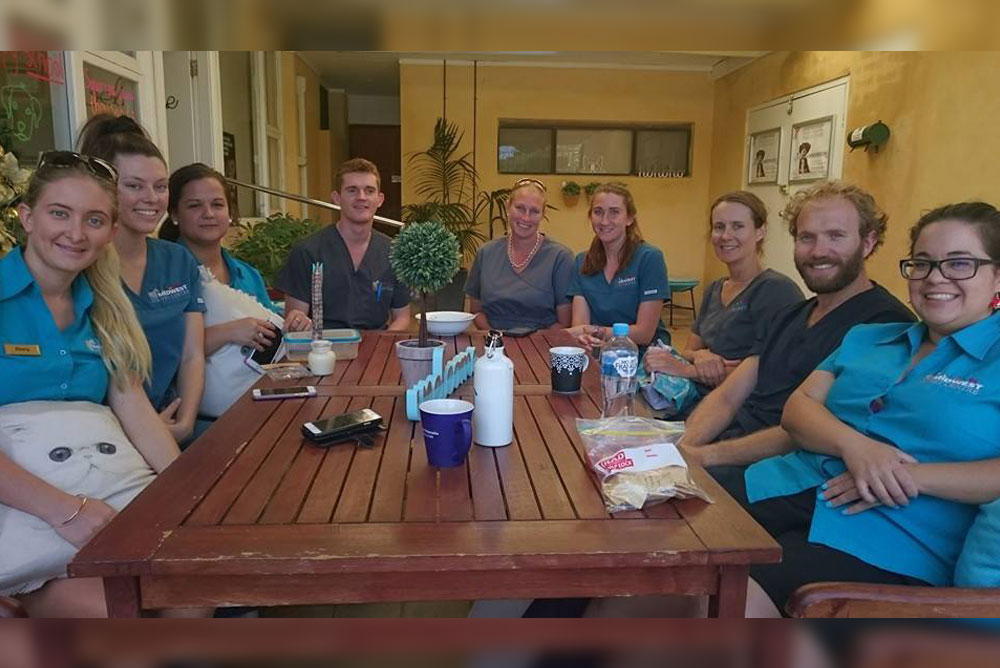 A group of eight people, five women and three men, sit around a wooden table in a relaxed outdoor setting. They have various items on the table, including cups and snacks. Some are smiling and they appear to be staff, possibly veterinarians or working in a healthcare setting.